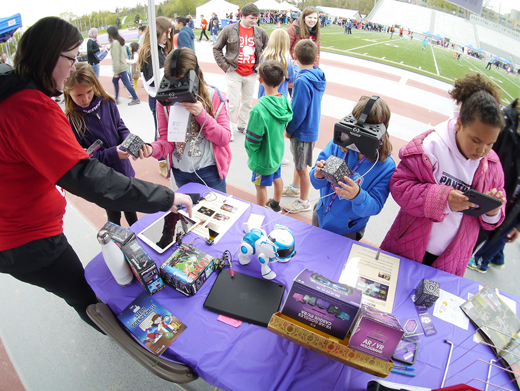 Children gather at technology booth, two wearing virtual reality headsets, one playing on a tablet, while a volunteer leads them through an activity
