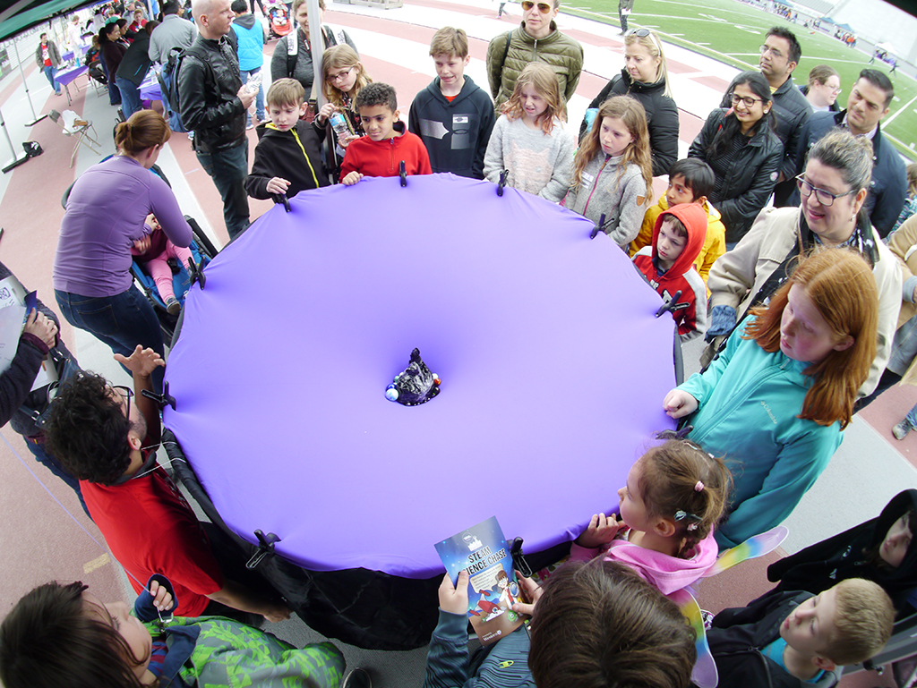 Children gather around a purple tarp stretched into a large circle. Marbles are gathered at the centre of the sheet.