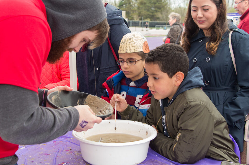 Visitors and booth in front of a bowl of murky brown water while volunteer sifts though sand looking for gold